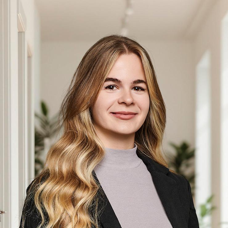 Junge Frau mit langen, blonden Haaren steht in einem hellen Flur mit Pflanzen im Hintergrund.