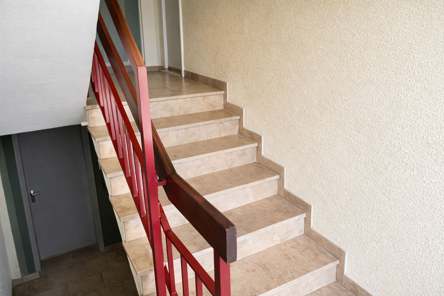 Treppe mit beigem Fliesenbelag und rotem Geländer in einem Wohnhausflur.