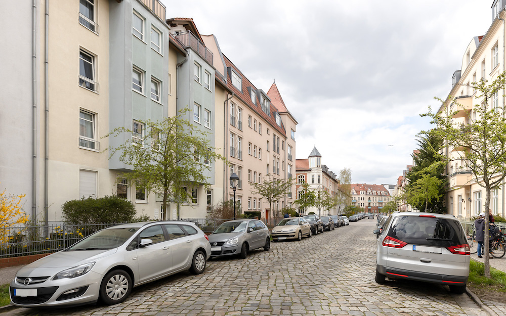 Wohnstraße mit mehrstöckigen Häusern, Autos am Straßenrand und kleinen Bäumen.