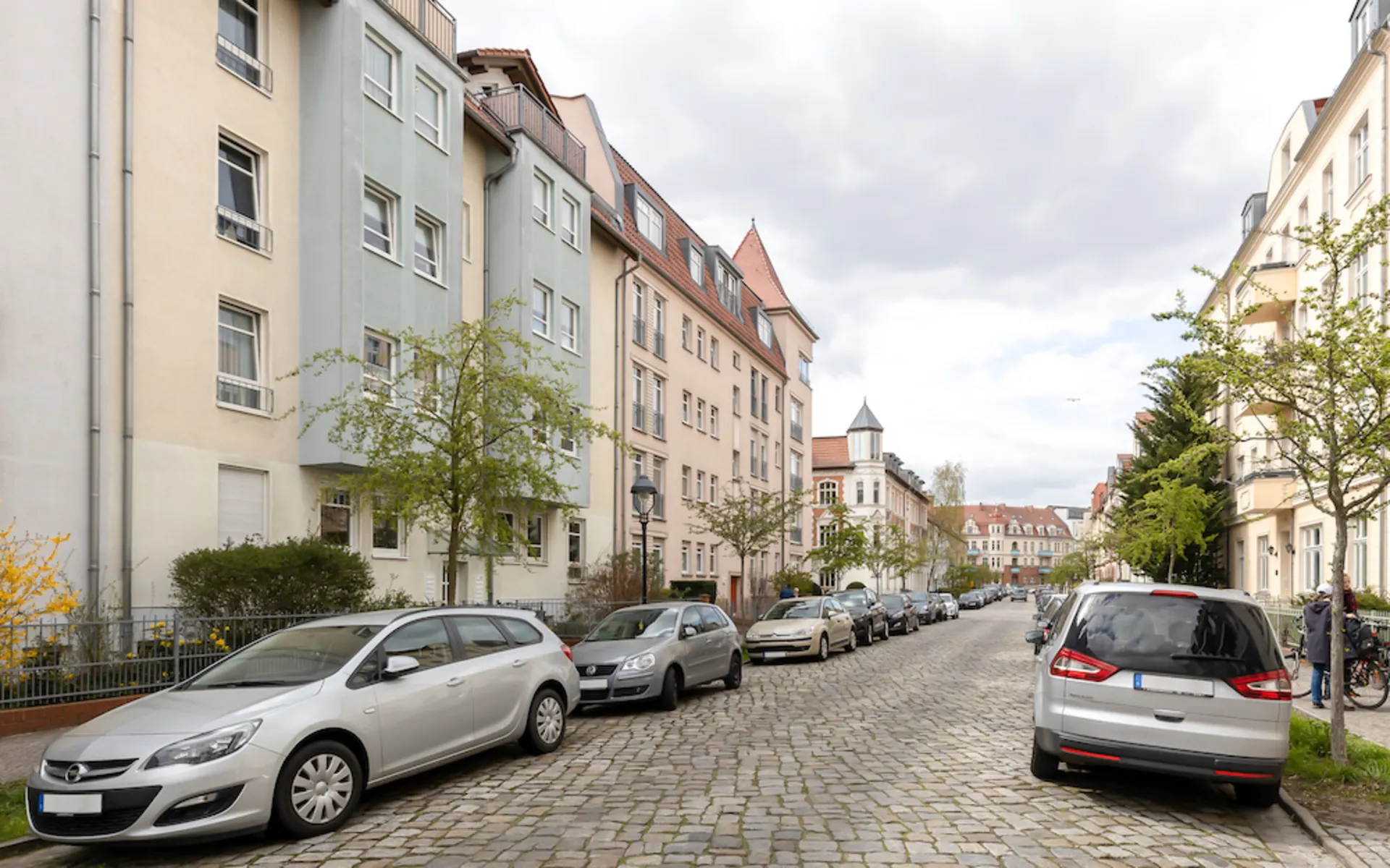 Wohnstraße mit mehrstöckigen Häusern, Autos am Straßenrand und kleinen Bäumen.