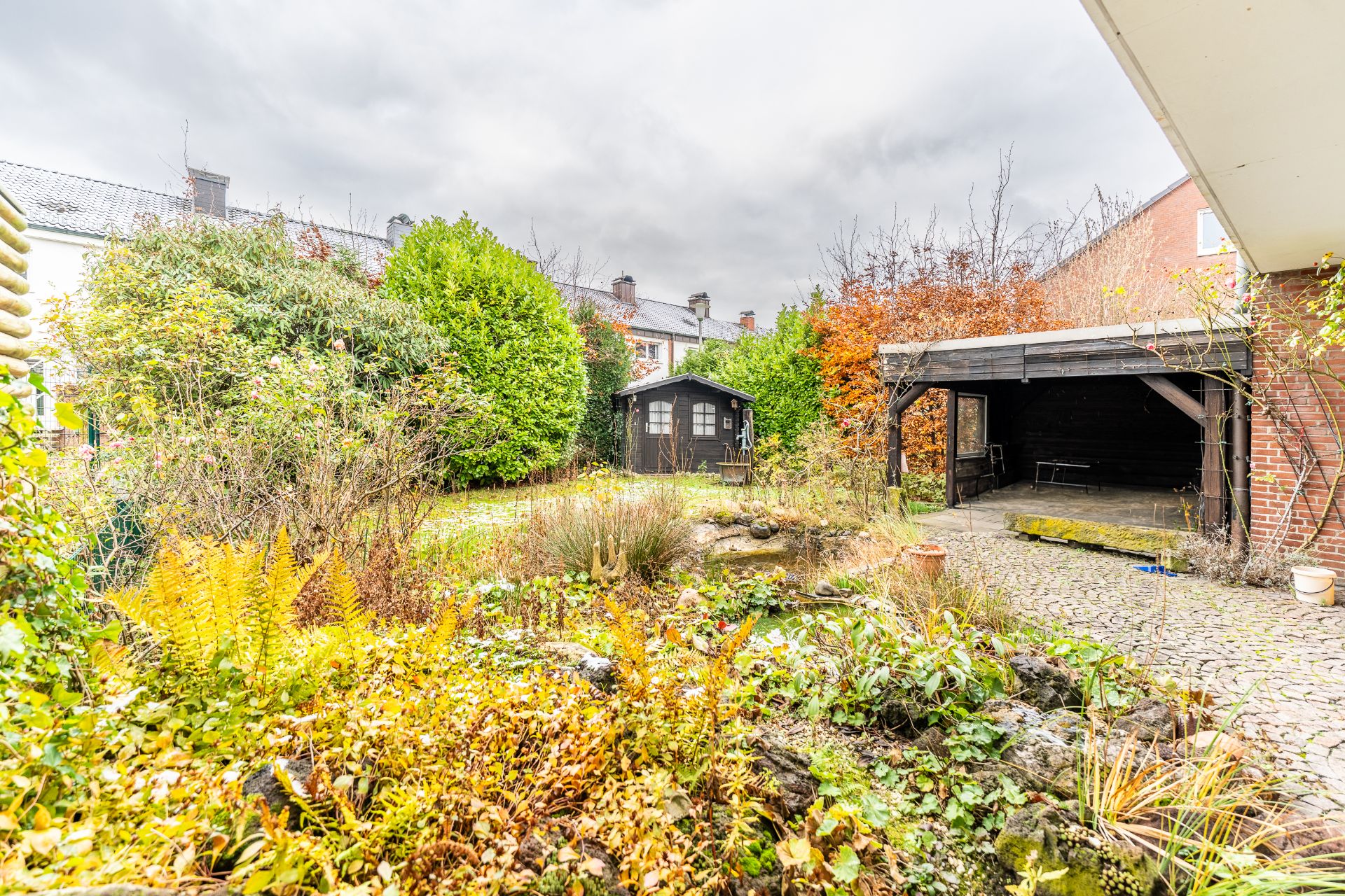 Garten mit Teich, vielen Pflanzen, einer Holzhütte und einer überdachten Terrasse.
