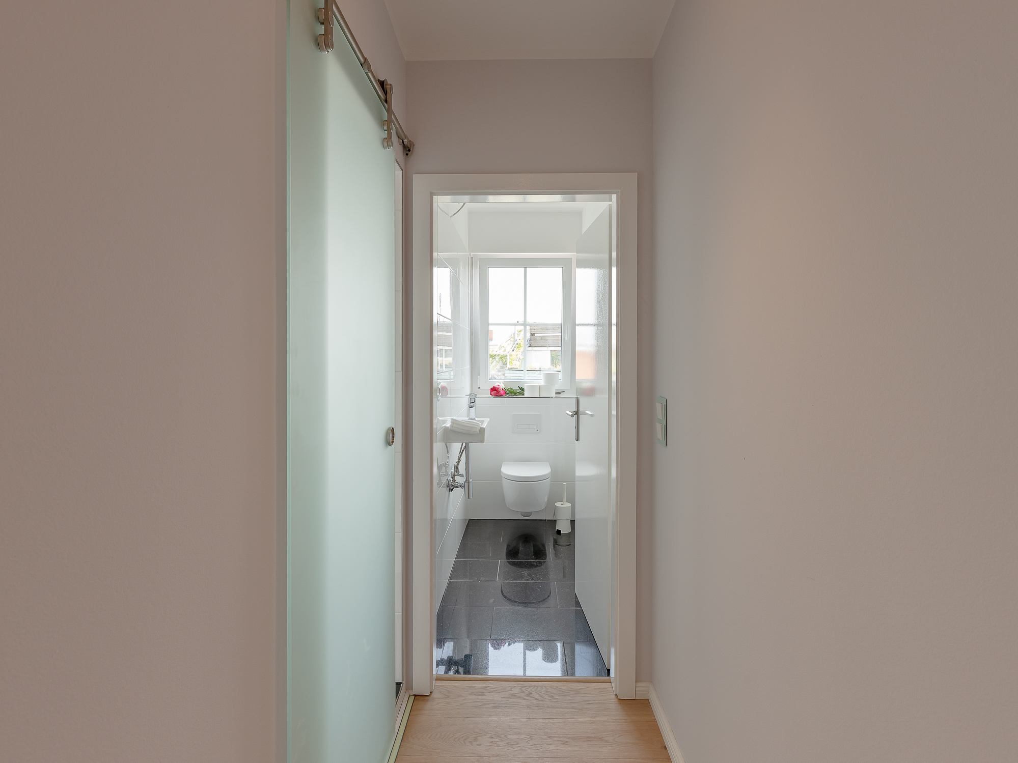 Blick durch einen Flur in ein helles Badezimmer mit WC, Waschbecken und Fenster.