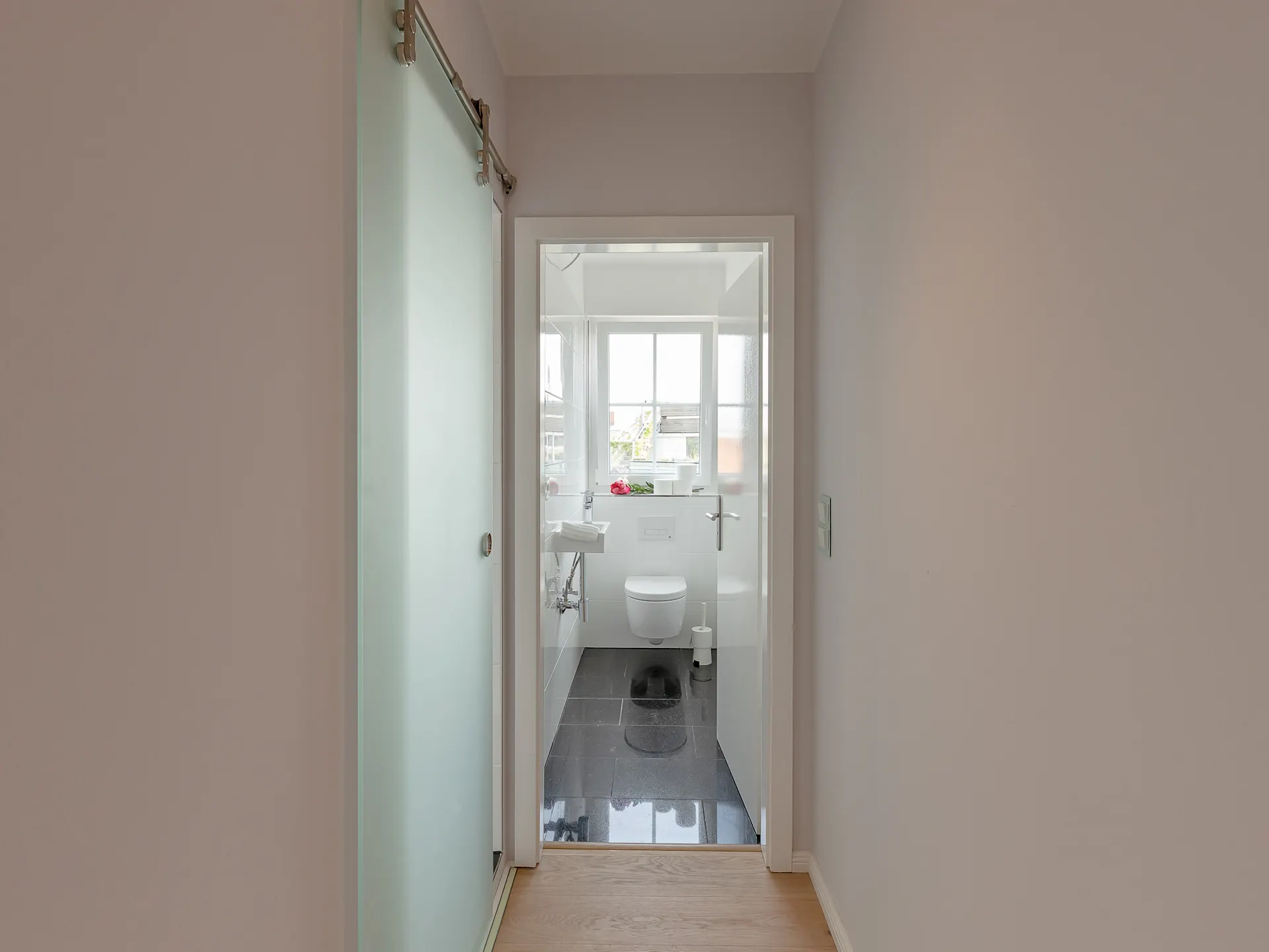 Blick durch einen Flur in ein helles Badezimmer mit WC, Waschbecken und Fenster.