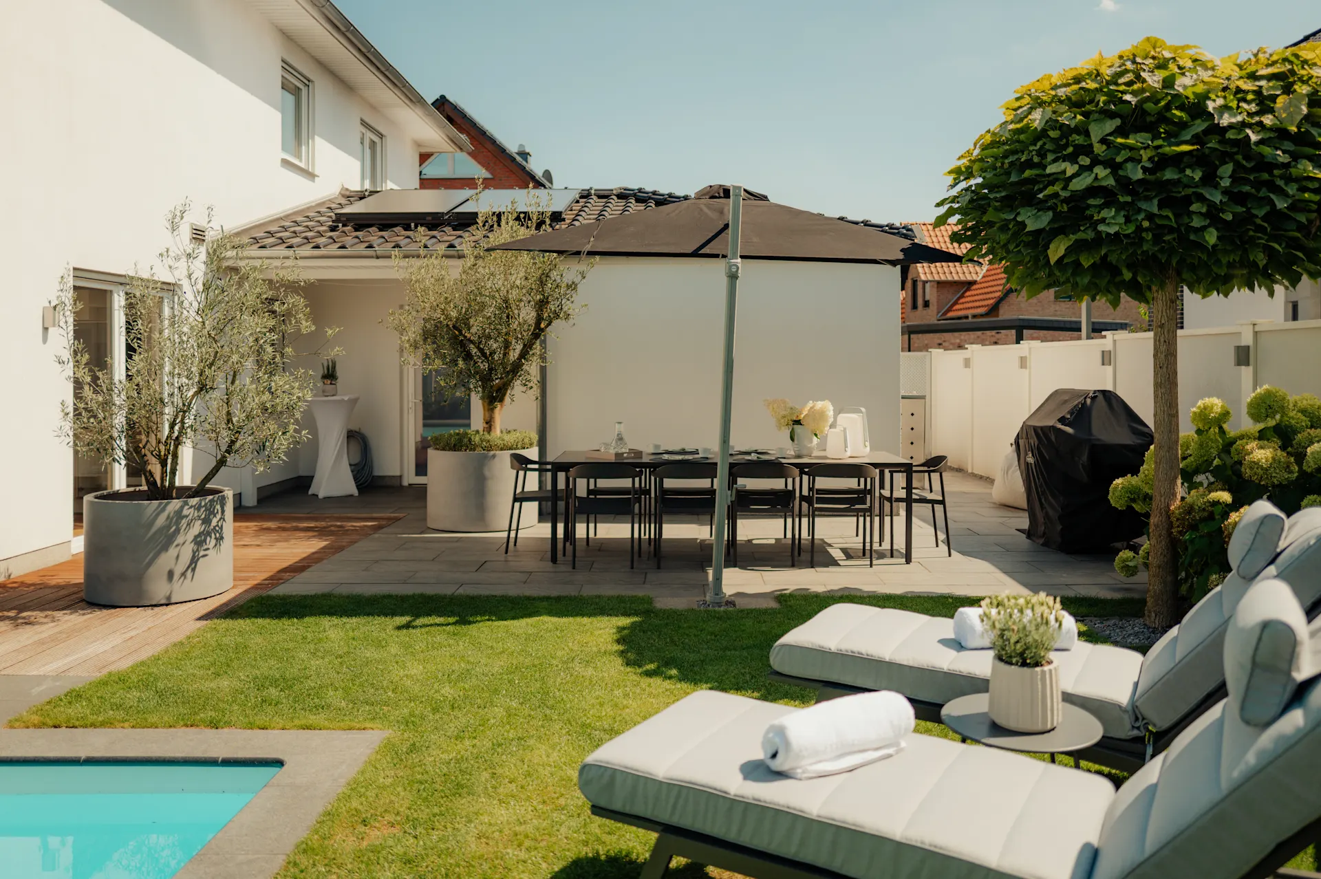 Garten mit Pool, Liegestühlen, Esstisch, Sonnenschirm und Grill neben einem modernen Wohnhaus.