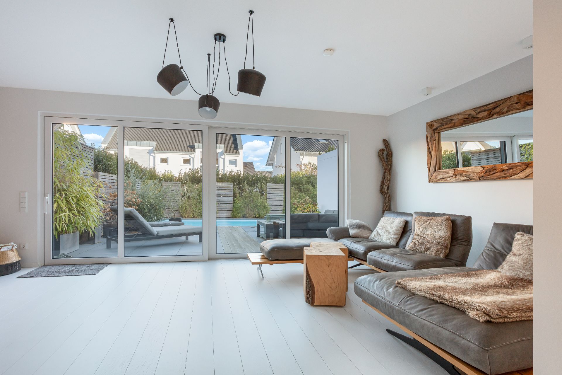 Modernes Wohnzimmer mit großen Fenstern, Blick auf Terrasse und Pool, gemütliche Sofas und dekorativem Spiegel.