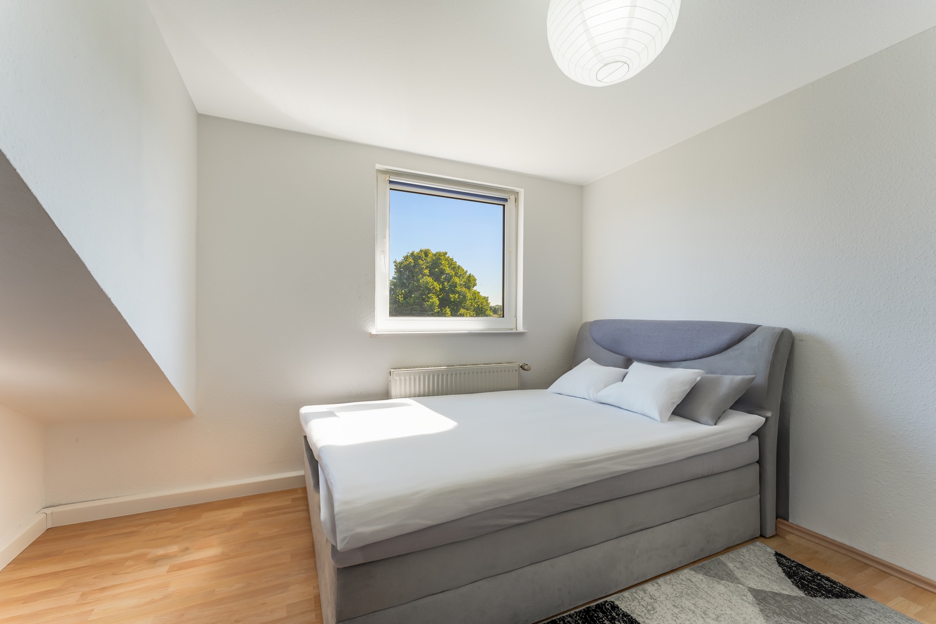 Helles Schlafzimmer mit Doppelbett, Fenster, hellen Wänden und Holzfußboden.