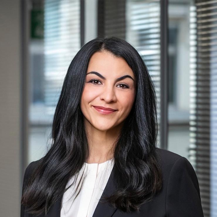 Eine Frau mit Brille und dunklem Blazer steht vor einem Fenster mit Jalousien.