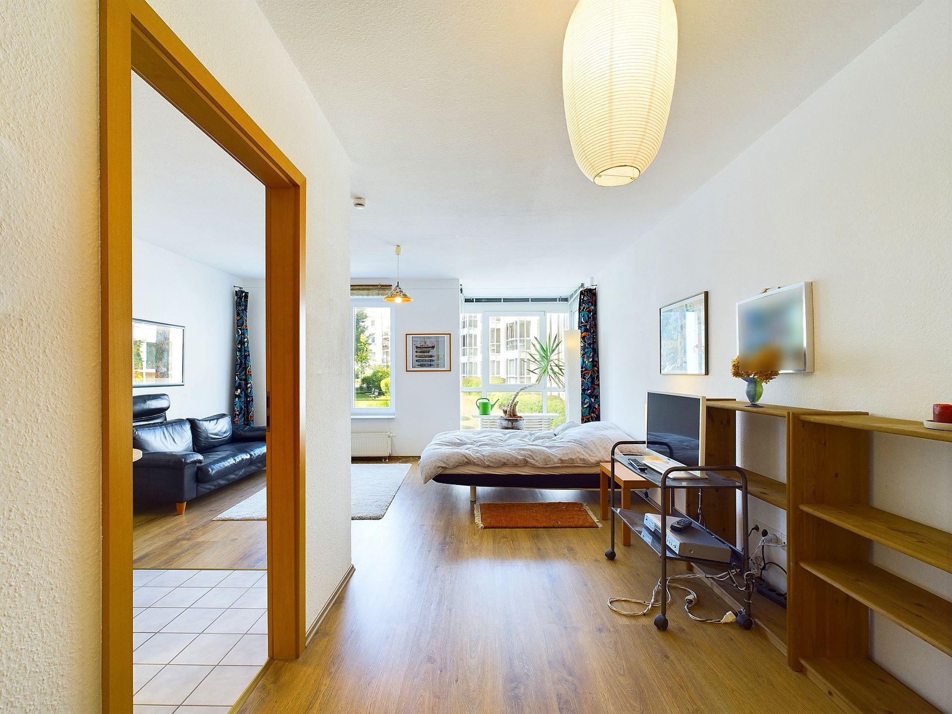 Helles Zimmer mit Bett, Fernseher, Regal und Blick ins Wohnzimmer mit Sofa.