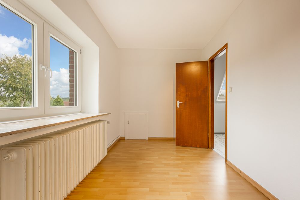 Helles Zimmer mit Holzfußboden, Fenster, Heizkörper und geöffneter Holztür.