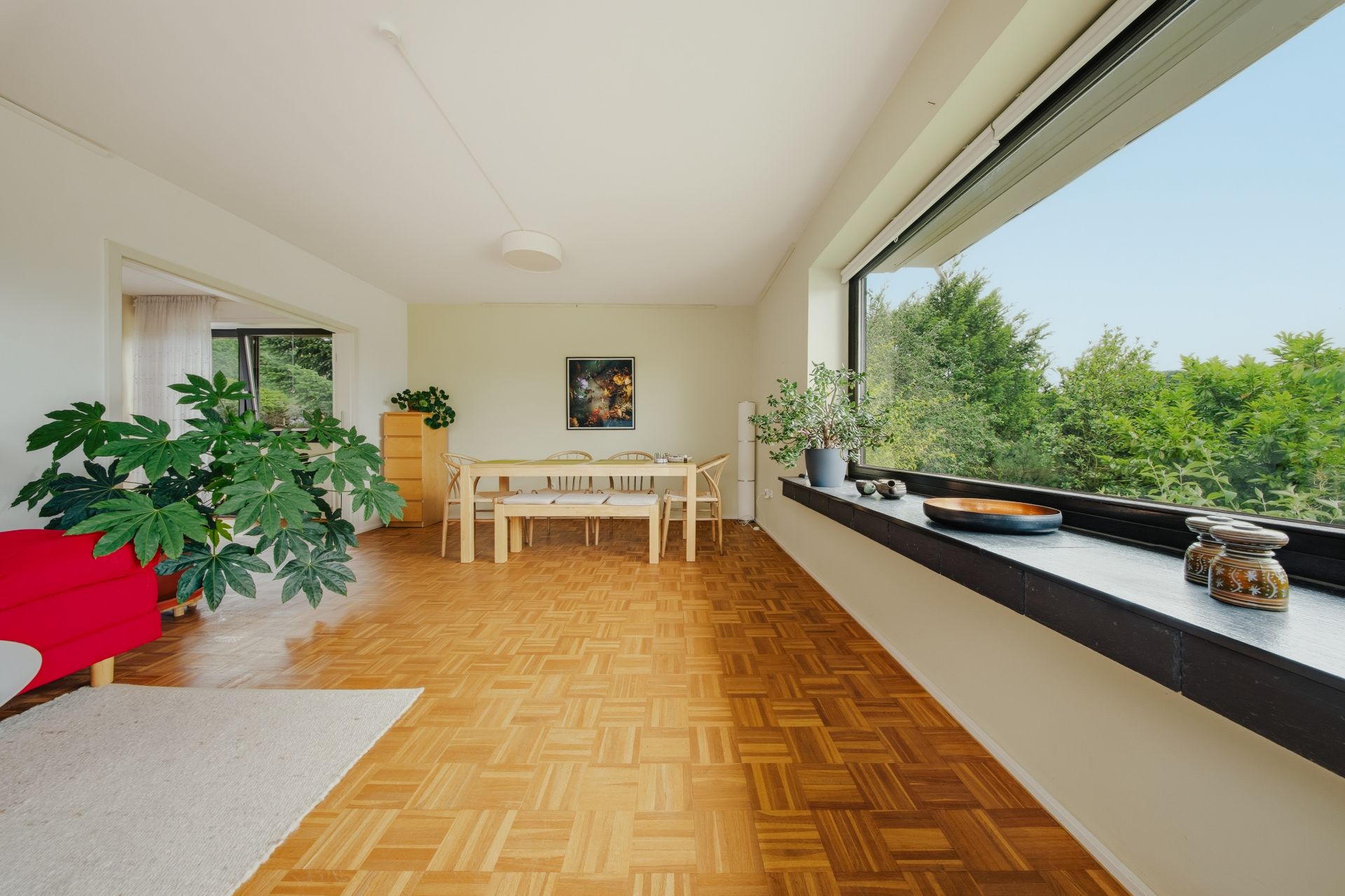 Helles Wohnzimmer mit großem Fenster, Holzboden, Esstisch und Zimmerpflanzen.