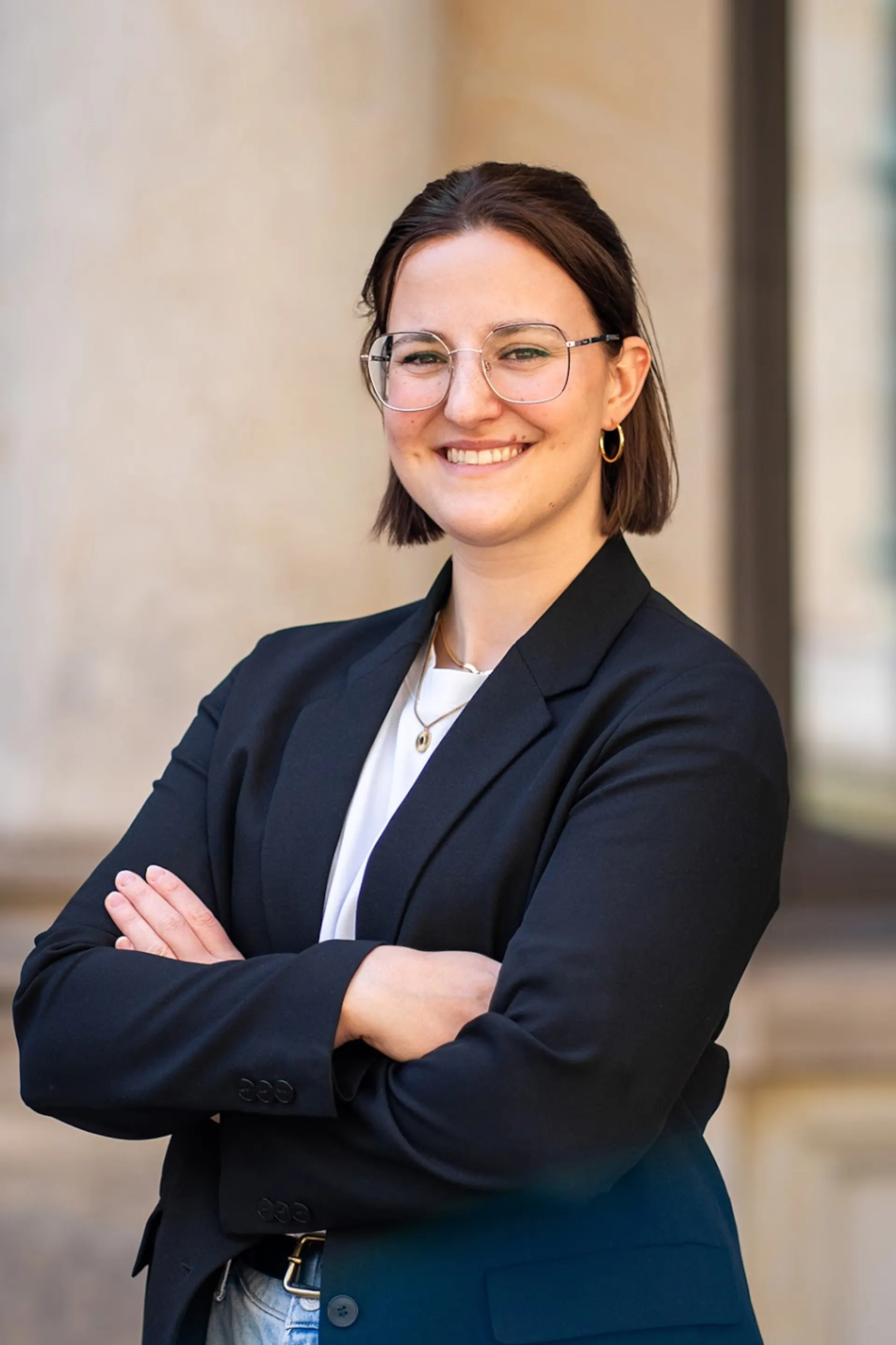 Eine lächelnde Person mit Brille und schwarzem Blazer steht mit verschränkten Armen vor einem Gebäude.