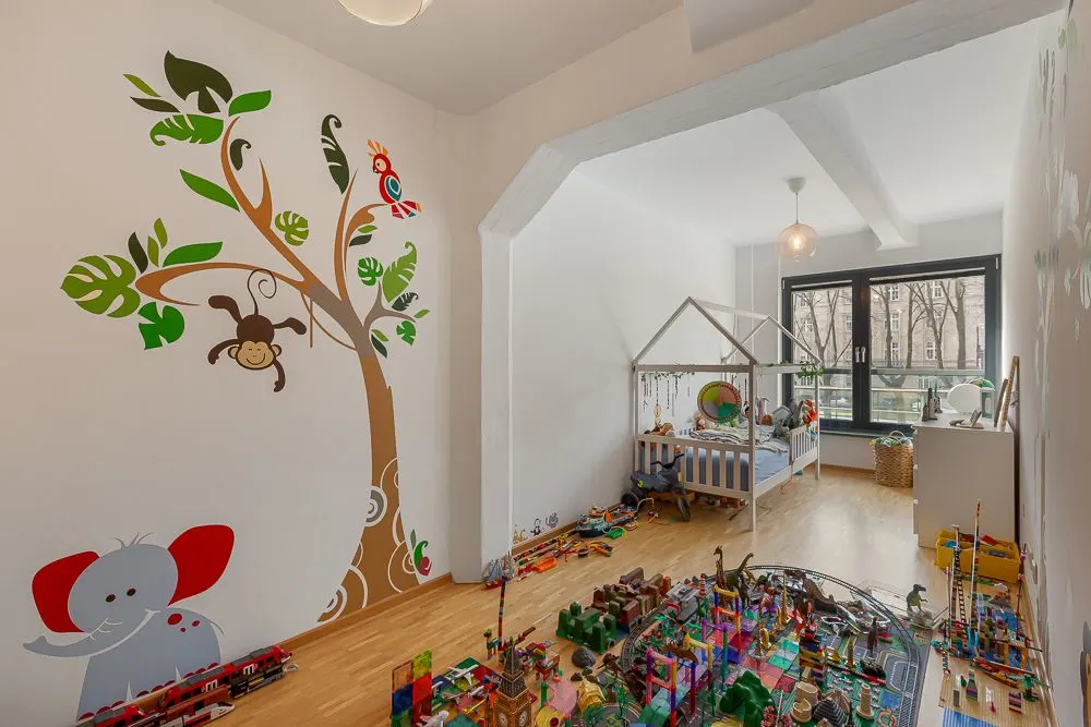 Kinderzimmer mit Wandaufklebern von Tieren, Spielzeug auf dem Boden und Kinderbett am Fenster.