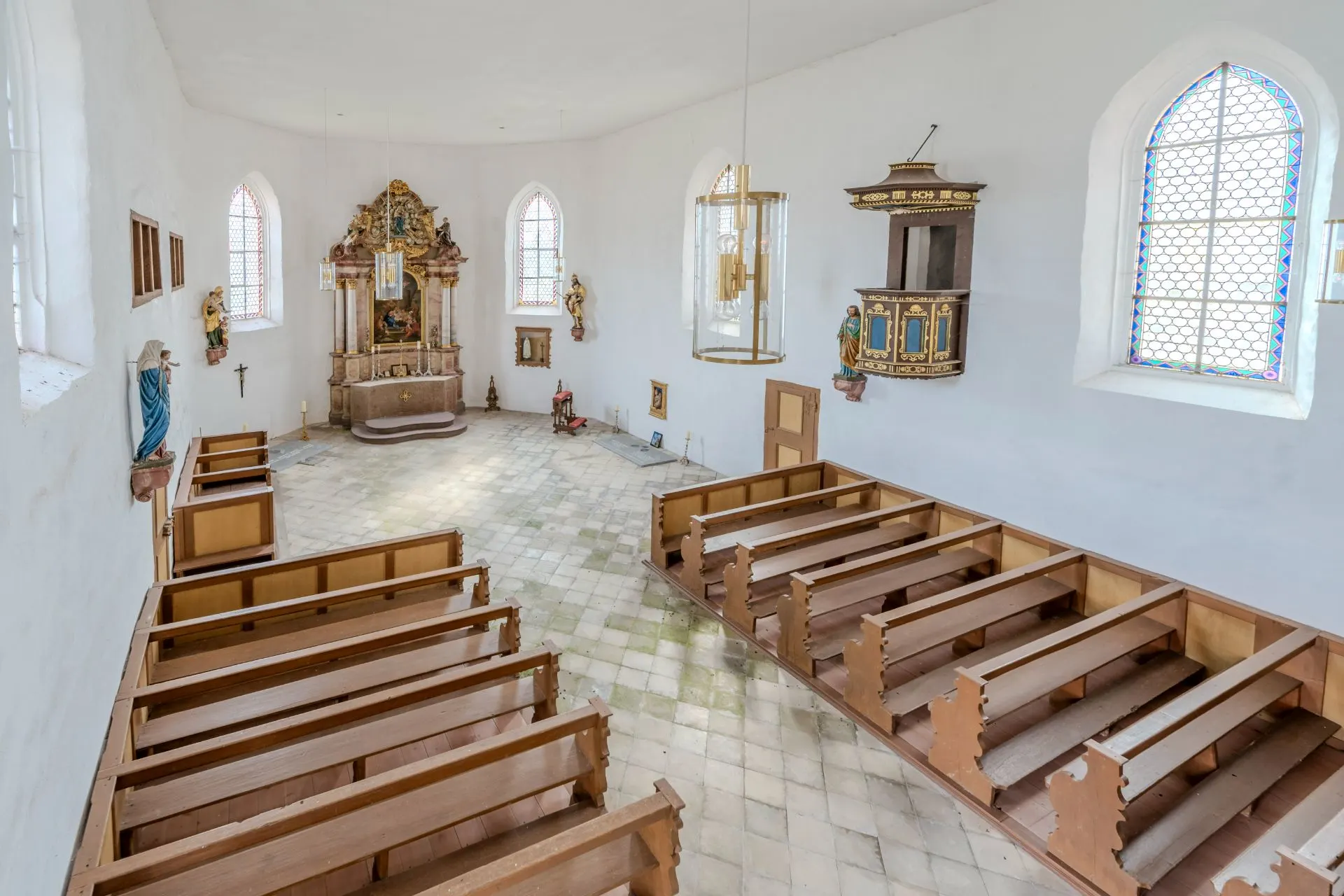 Innenraum einer kleinen Kirche mit Holzbänken, Altar und bunten Fenstern.