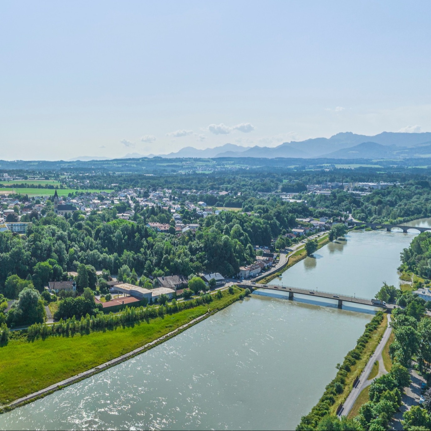 Panoramaaufnahme mit Fluss, Brücke, grünen Wiesen und Wohnhäusern im Hintergrund.