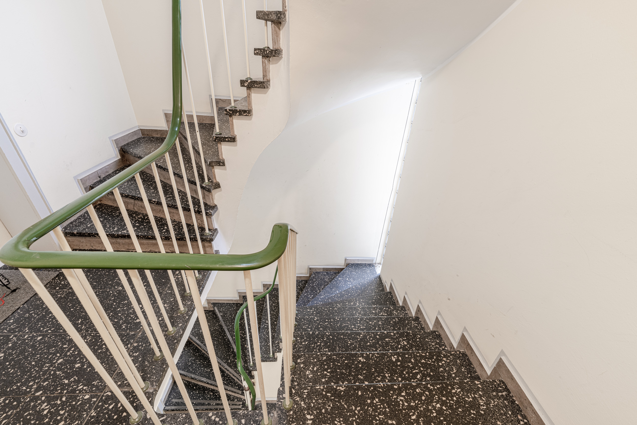 Treppenhaus mit grünem Handlauf und Fenster, Tageslicht fällt auf die Stufen.