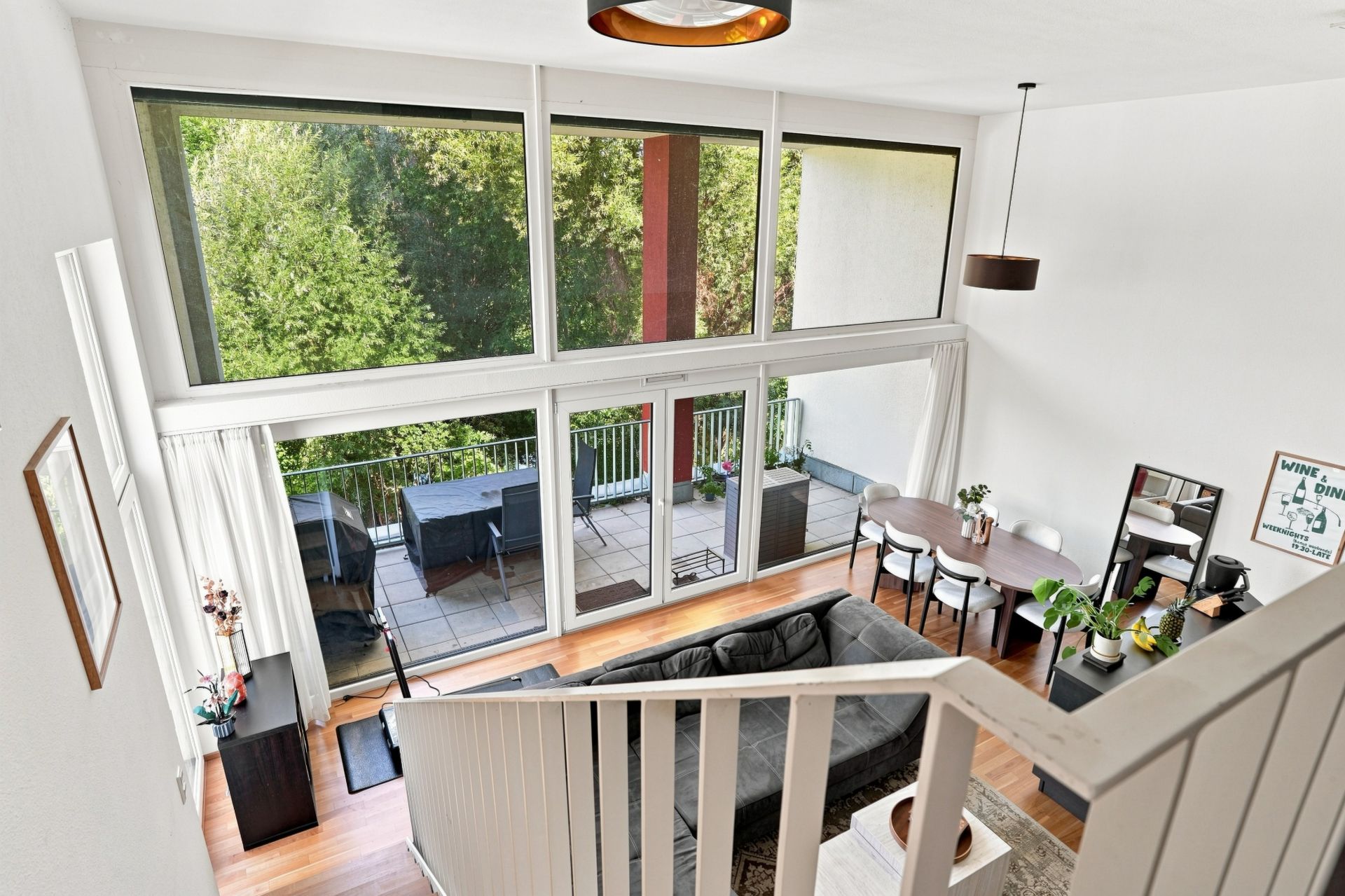 Helles Wohnzimmer mit großer Fensterfront, Sofa, Esstisch und Zugang zu Balkon mit Gartenblick.