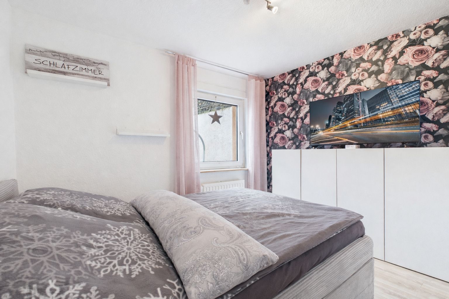 Schlafzimmer mit Doppelbett, floraler Tapete, Fernseher und Fenster mit rosa Gardine.