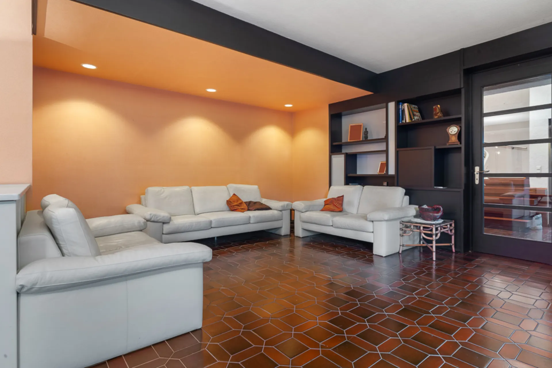 Wohnzimmer mit drei hellen Sofas, braunem Fliesenboden und eingebauten Regalen an der Wand.