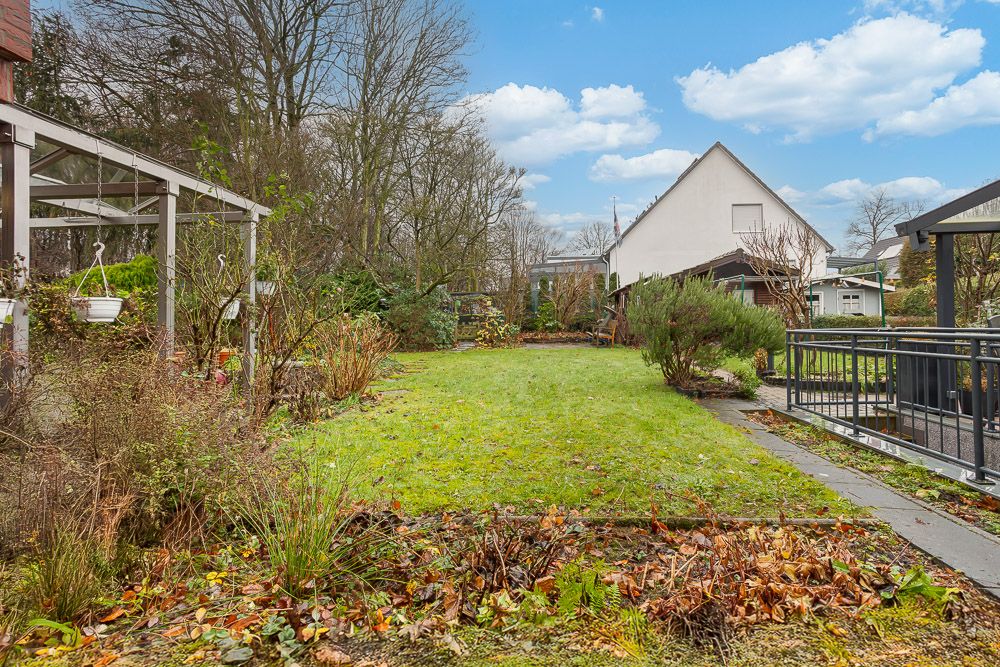 Garten mit Rasenfläche, Pflanzen und überdachter Terrasse neben einem Wohnhaus.