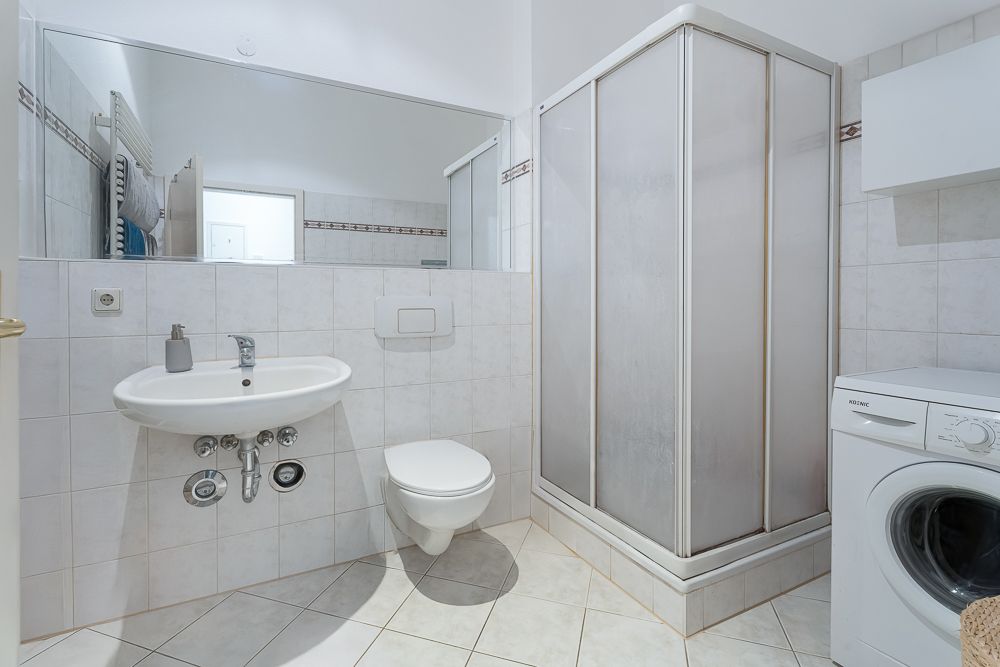 Helles Badezimmer mit Dusche, Waschbecken, WC, großem Spiegel und Waschmaschine.