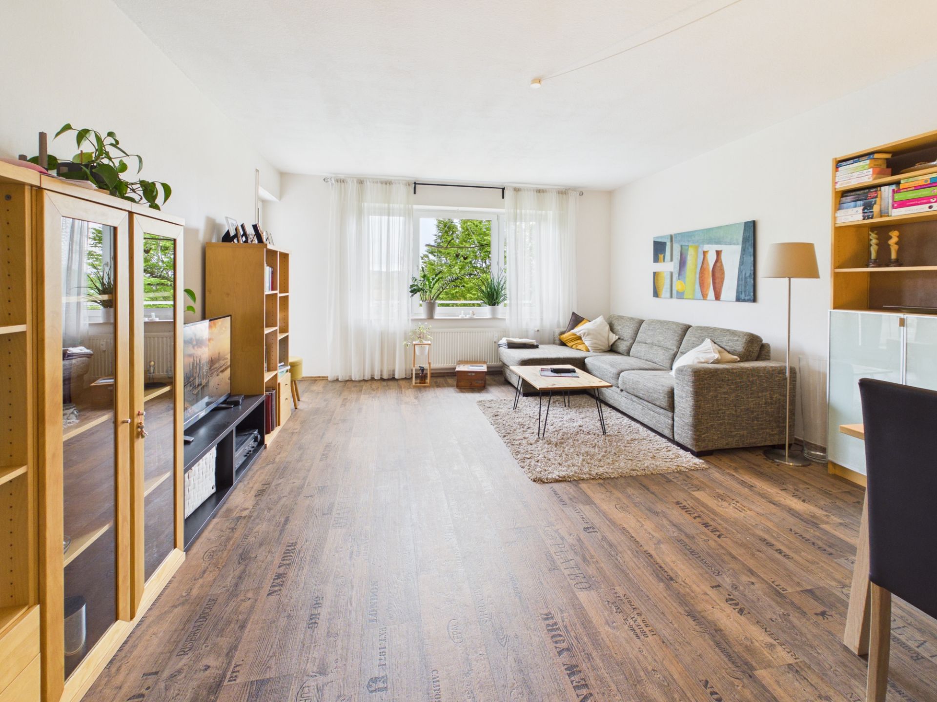 Helles Wohnzimmer mit Holzfußboden, grauem Sofa, Regalen und einem großen Fenster mit weißen Vorhängen.