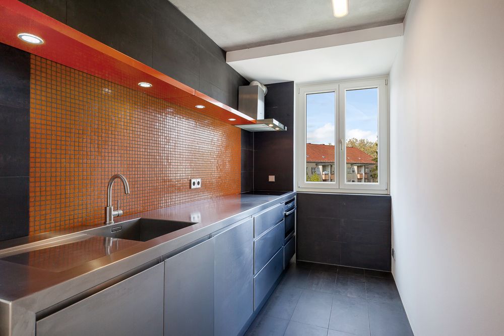 Moderne Küche mit Edelstahlarbeitsfläche, orangefarbener Fliesenwand und Fenster.