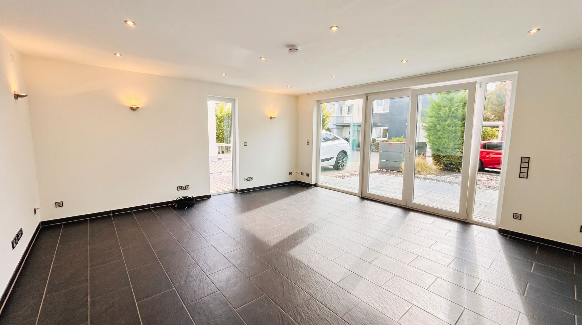 Leeres Wohnzimmer mit dunklem Fliesenboden und großen Fenstern zur Terrasse.