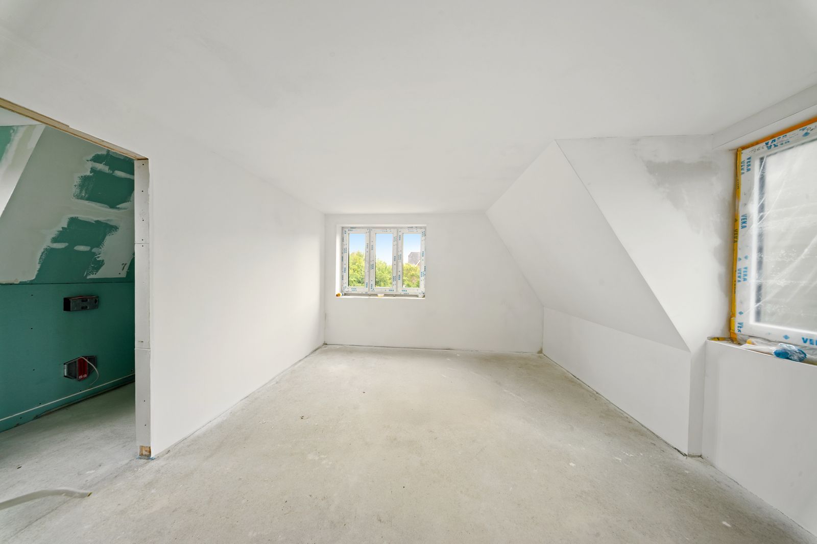 Leerer, heller Raum mit weißen Wänden, Fenster in der Mitte und Baustellenmaterial am Rand.