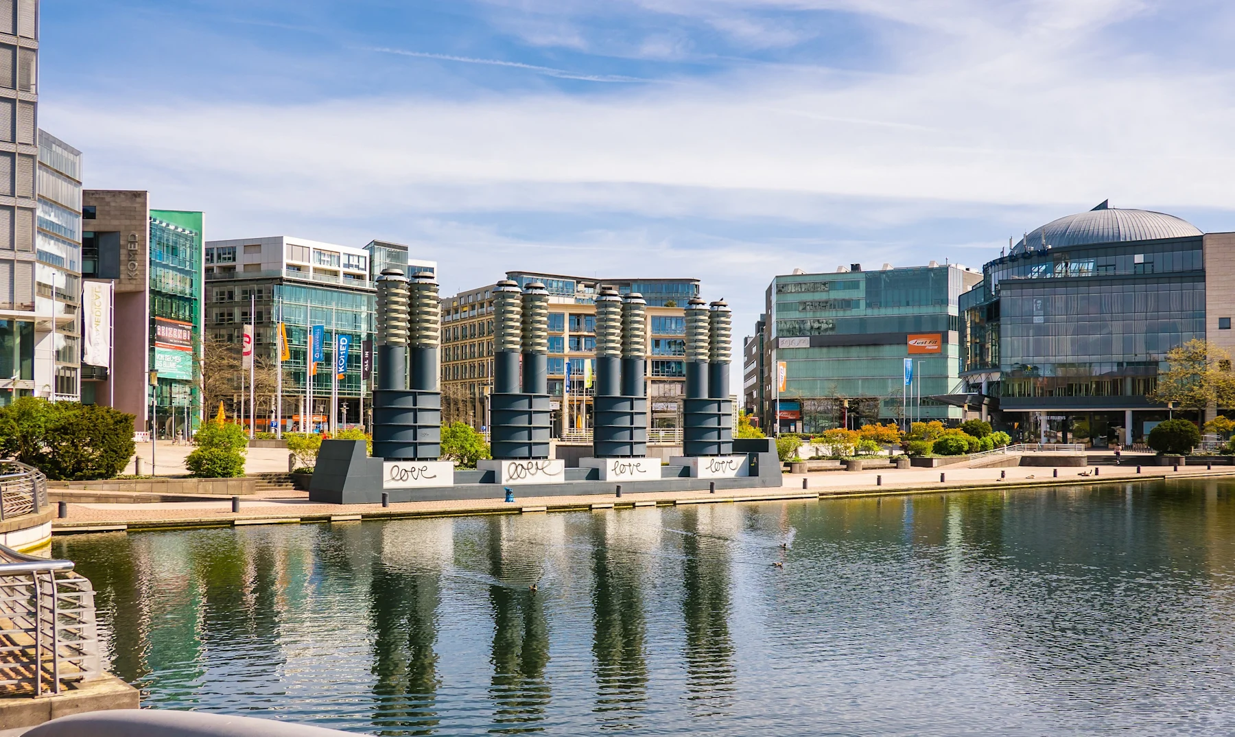 Moderne Bürogebäude und Kunstinstallation an einem Wasserbecken bei sonnigem Wetter.