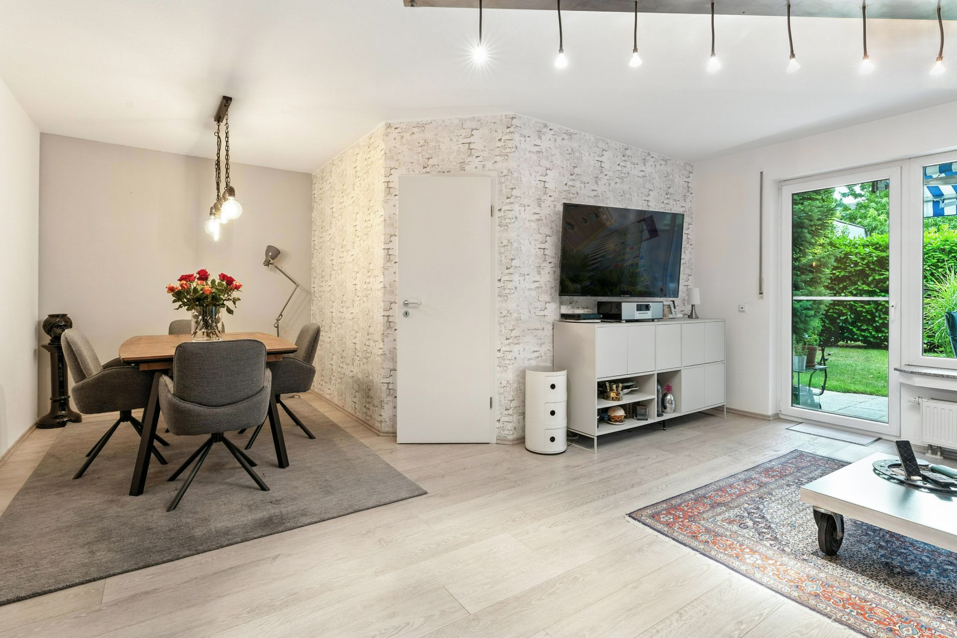 Modernes Wohnzimmer mit Essbereich, Fernseher, Glastür zum Garten und hellen Holzböden.