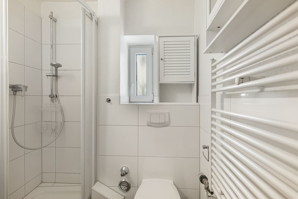 Kleines Badezimmer mit Dusche, Toilette, Heizkörper und kleinem Fenster.