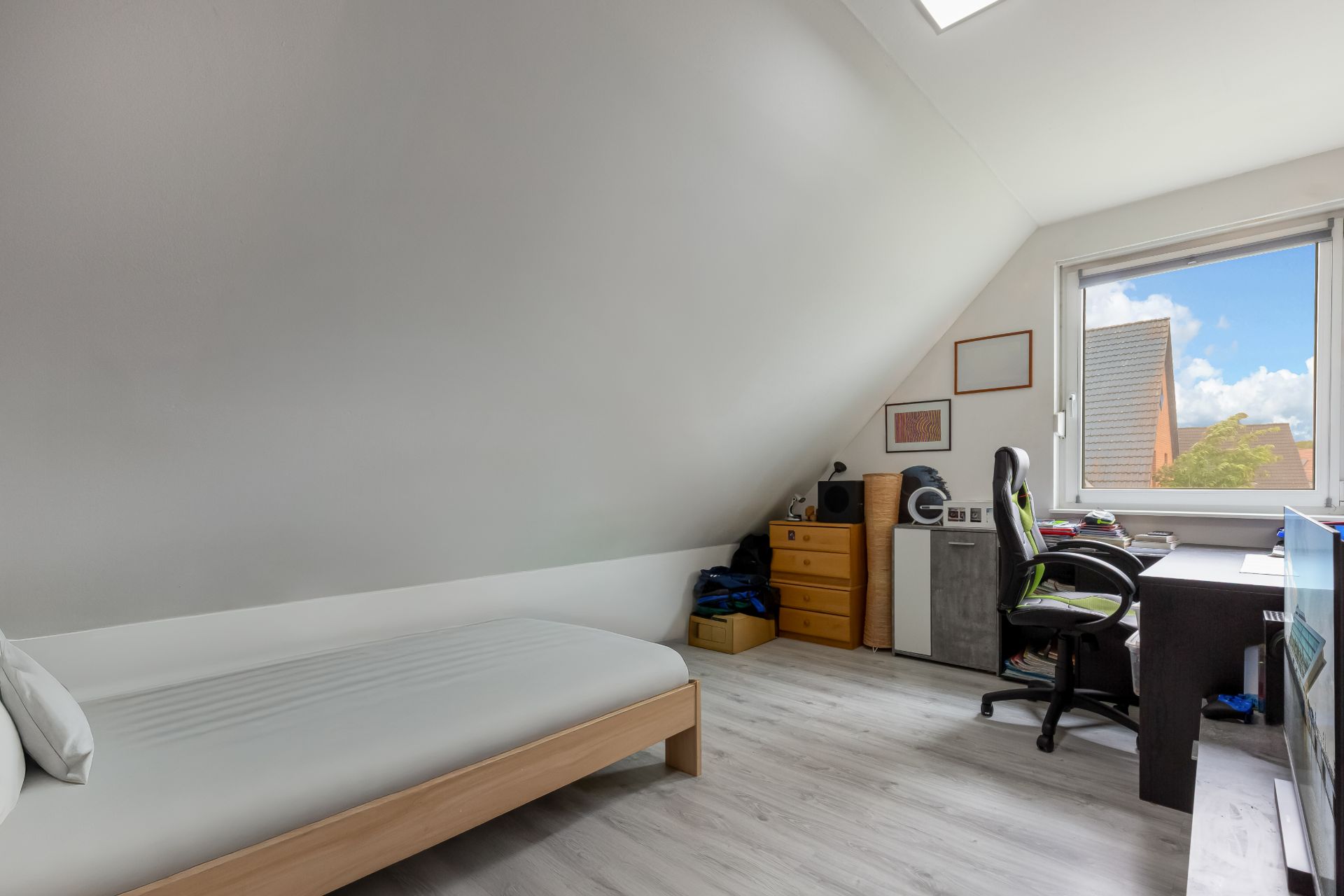 Helles Zimmer mit Einzelbett, Schreibtisch und Stuhl unter einer Dachschräge, Fenster mit Blick nach draußen.