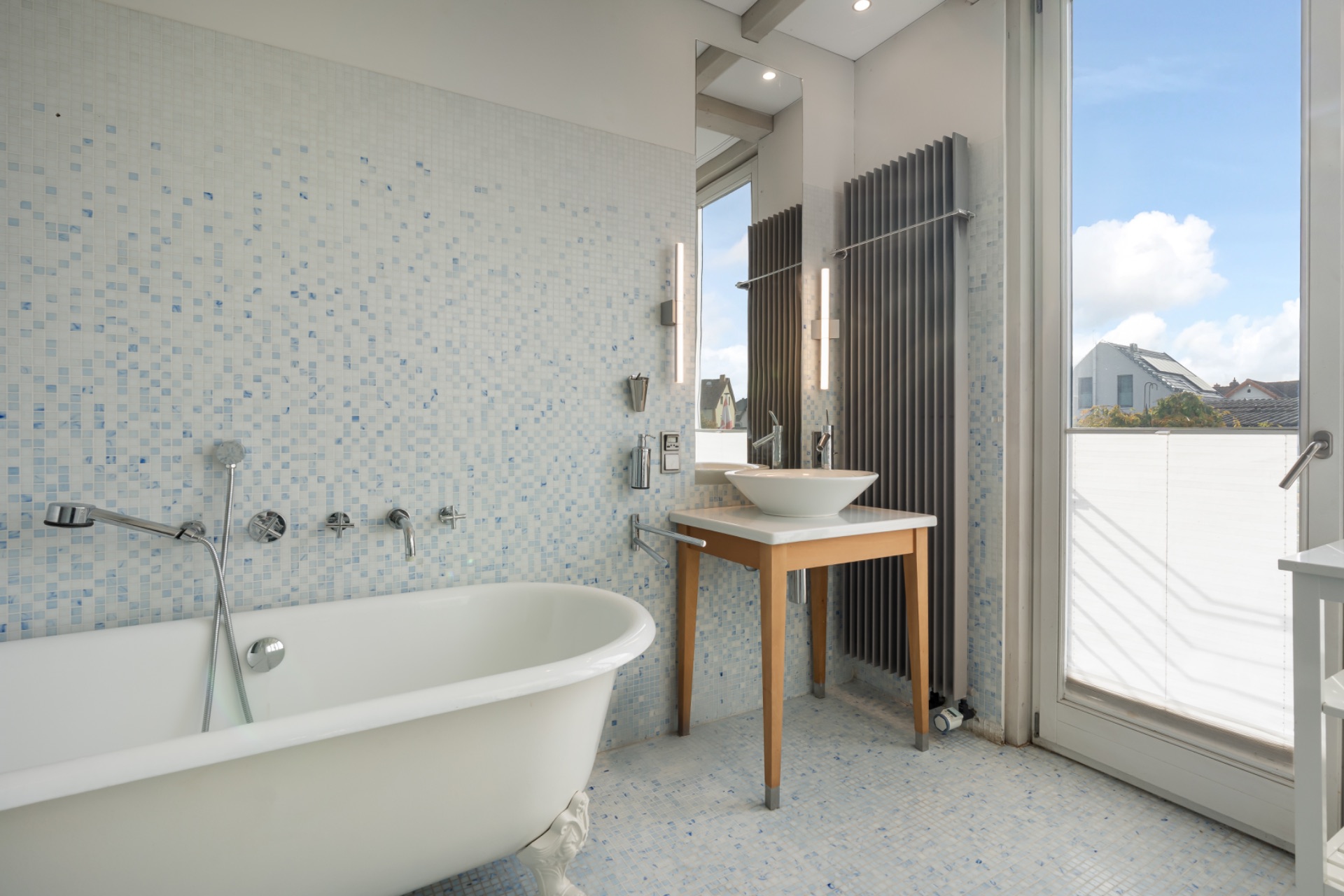 Helles Badezimmer mit freistehender Badewanne, Waschbecken auf Holztisch und Zugang zum Balkon.