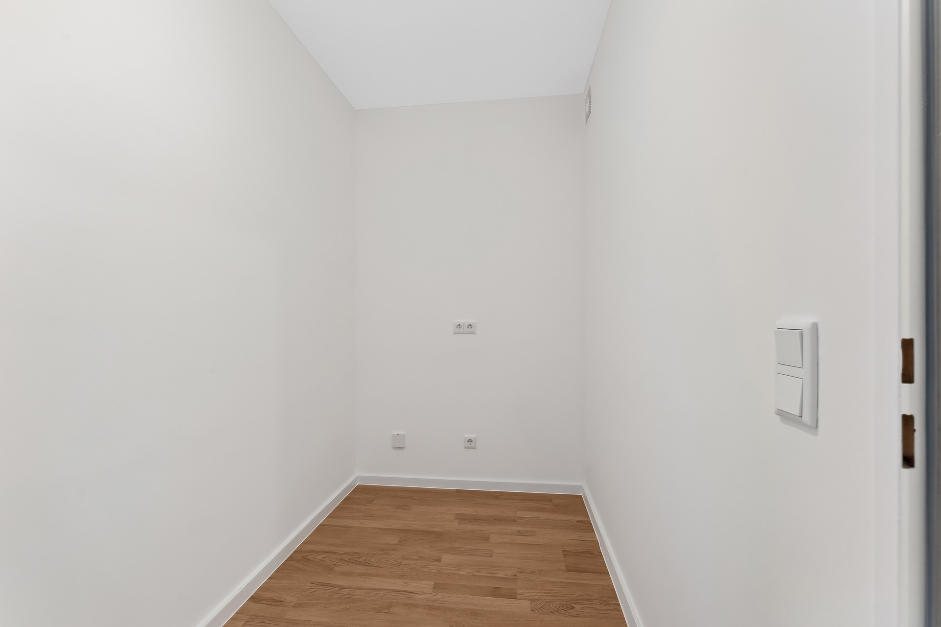 Kleiner, leerer Raum mit weißen Wänden und Holzboden, Steckdosen an der Wand.