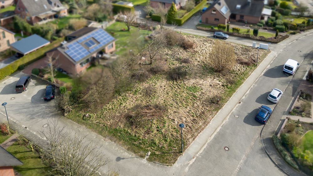 Luftaufnahme eines unbebauten, bewachsenen Grundstücks an einer Straßenecke mit geparkten Autos.