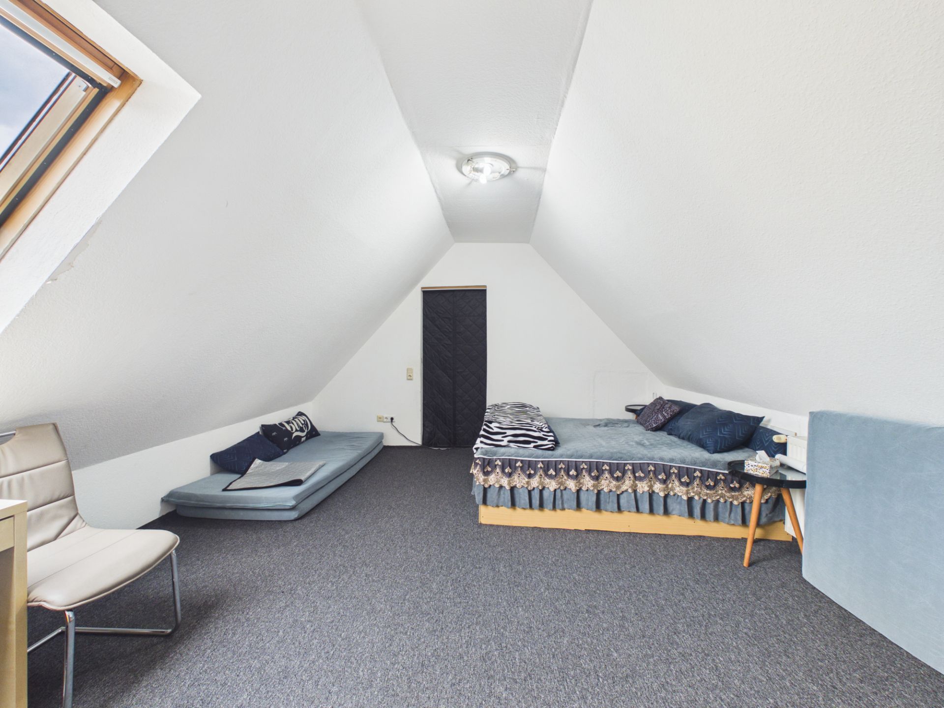 Dachzimmer mit zwei Betten, einem Stuhl, Dachfenster und Nachttisch.