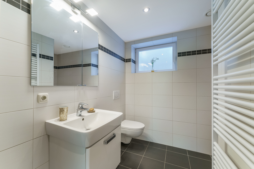 Modernes Badezimmer mit Waschbecken, Spiegel, Toilette und Fenster, weiße Fliesen an den Wänden.
