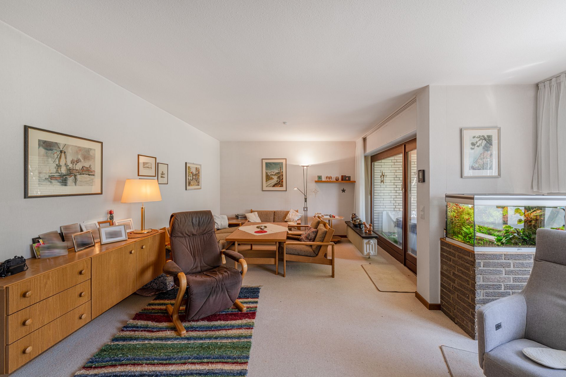 Helles Wohnzimmer mit Sesseln, Sofa, Aquarium, Teppich, Sideboard und Bildern an der Wand.