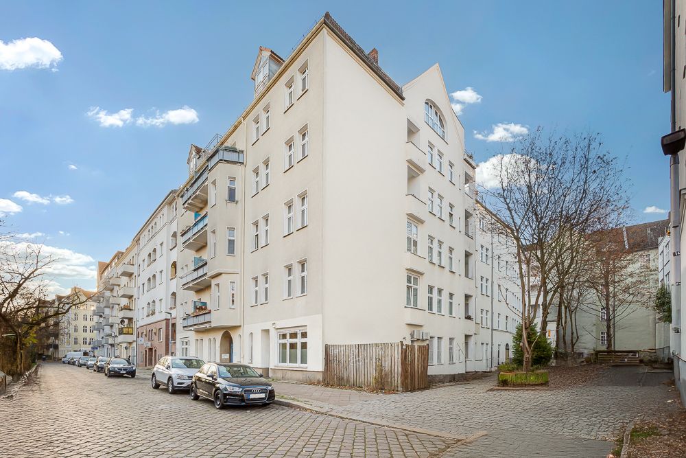 Eckiges Wohnhaus mit Balkonen an einer gepflasterten Straße, mehrere parkende Autos davor.