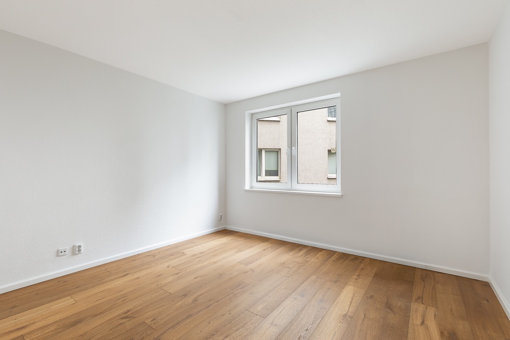Heller, leerer Raum mit Holzboden und einem Fenster, weiße Wände.