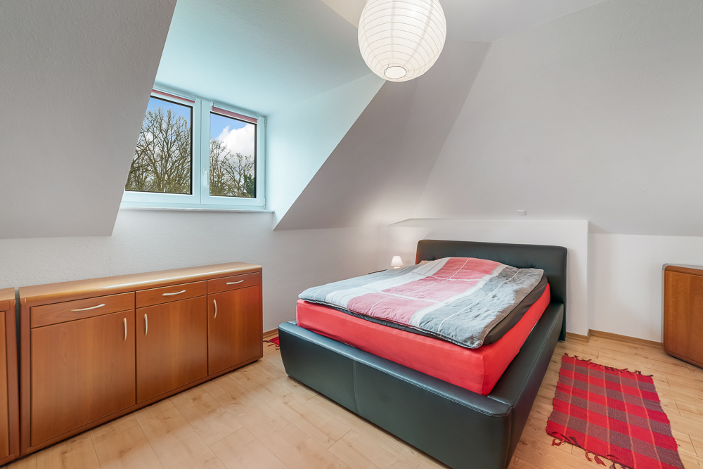 Schlafzimmer mit Doppelbett, Kommode, rotem Teppich und Fenster mit Blick auf Bäume.
