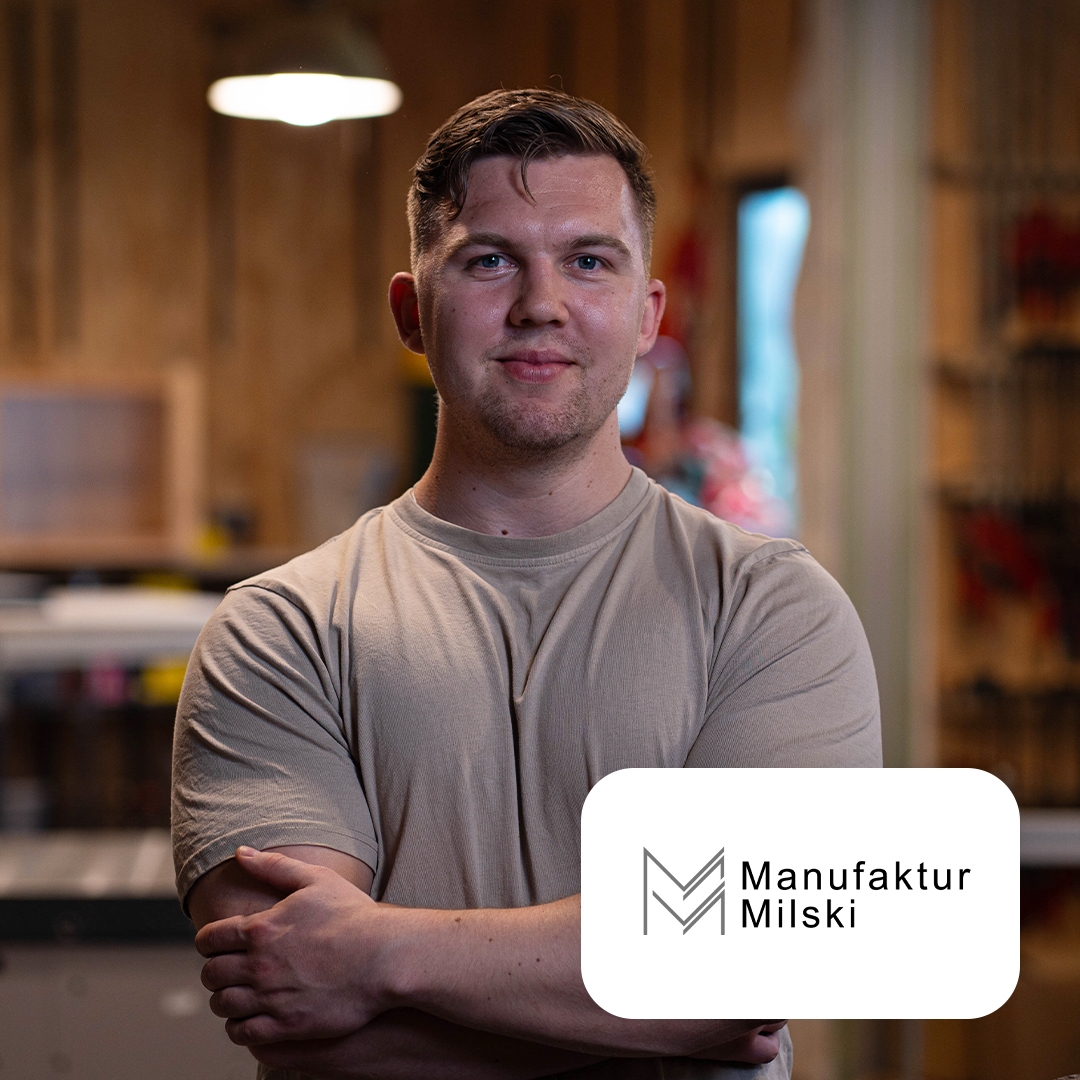 Ein Mann steht mit verschränkten Armen in einer Werkstatt. Logo mit Text: Manufaktur Milski unten rechts.