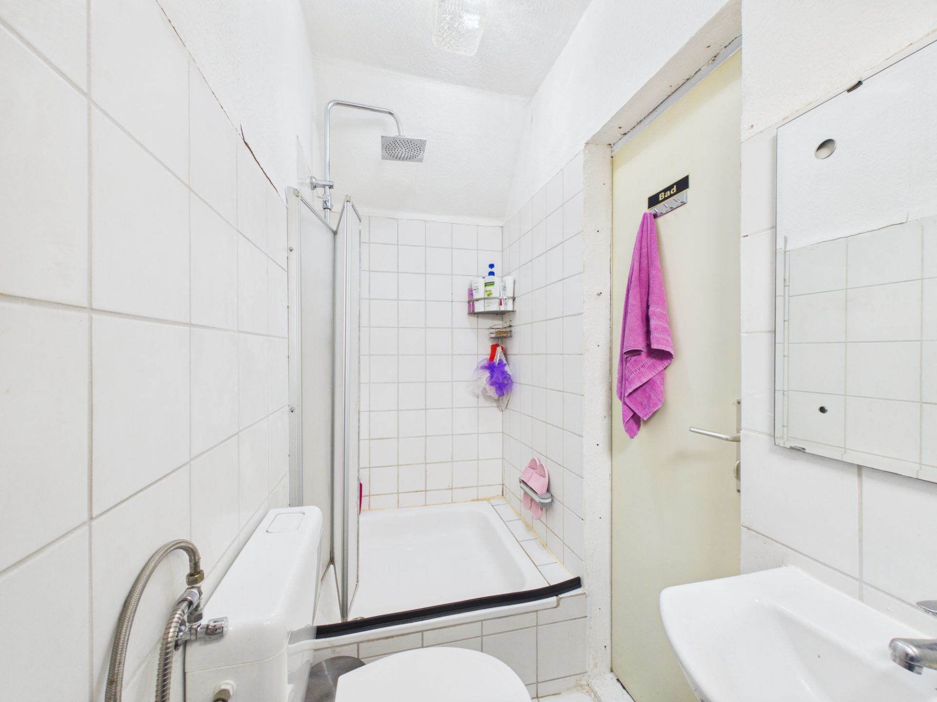 Kleines Badezimmer mit Dusche, Toilette, Waschbecken und Spiegel, weiß gefliest.
