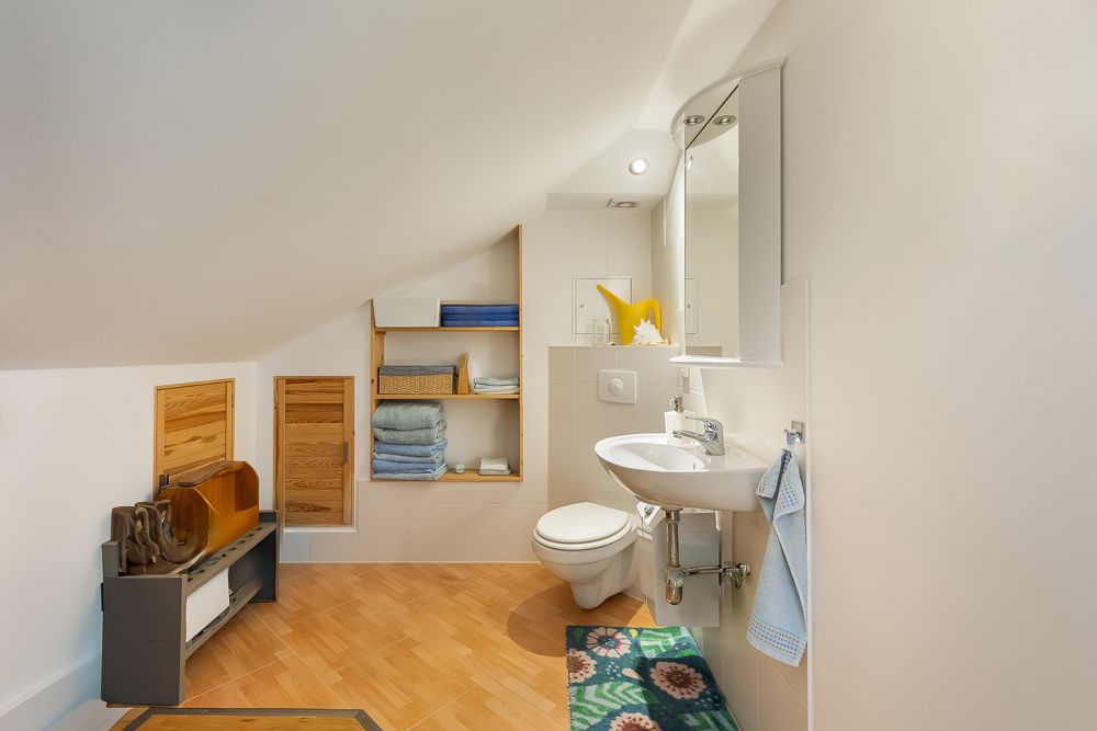 Helles Badezimmer mit Dachschräge, Toilette, Waschbecken, Regal mit Handtüchern und Spiegel.