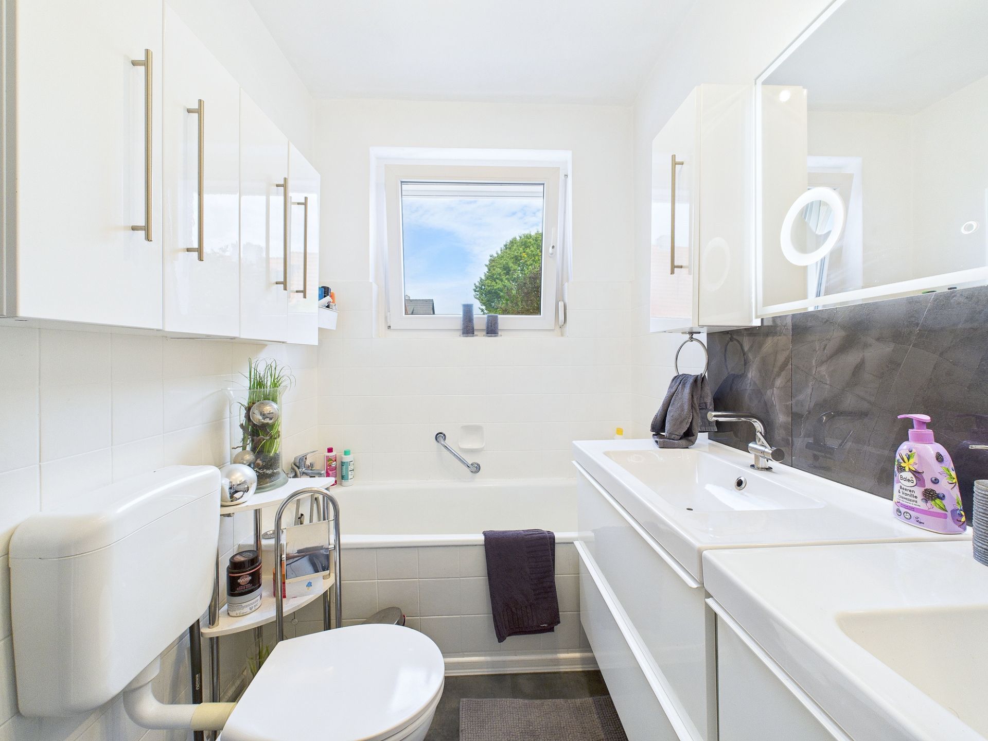 Helles Badezimmer mit Fenster, Waschbecken, Toilette, Badewanne und weißen Schränken.