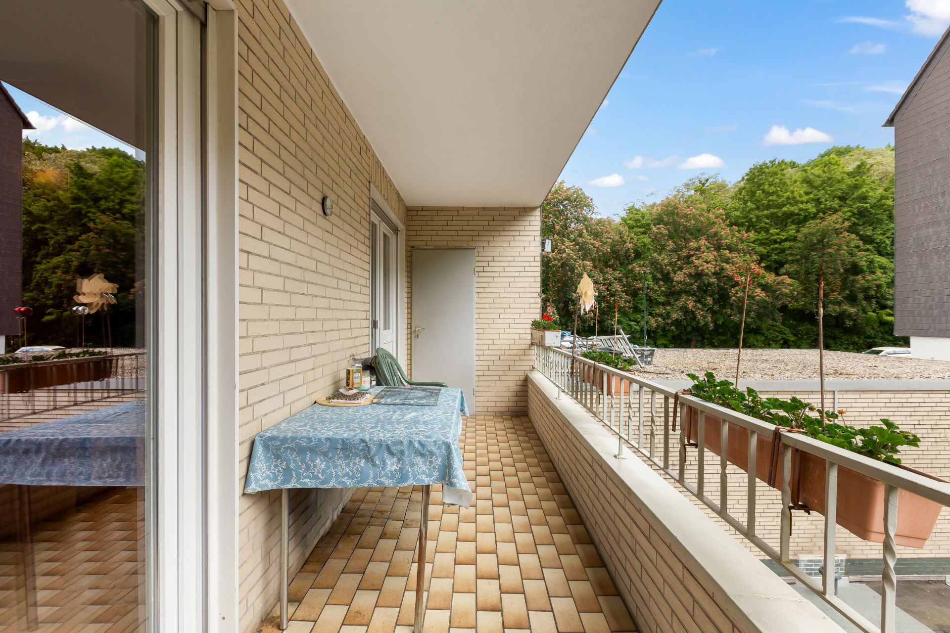 Balkon mit Tisch und Stühlen, Fliesenboden und Blick auf grüne Bäume.