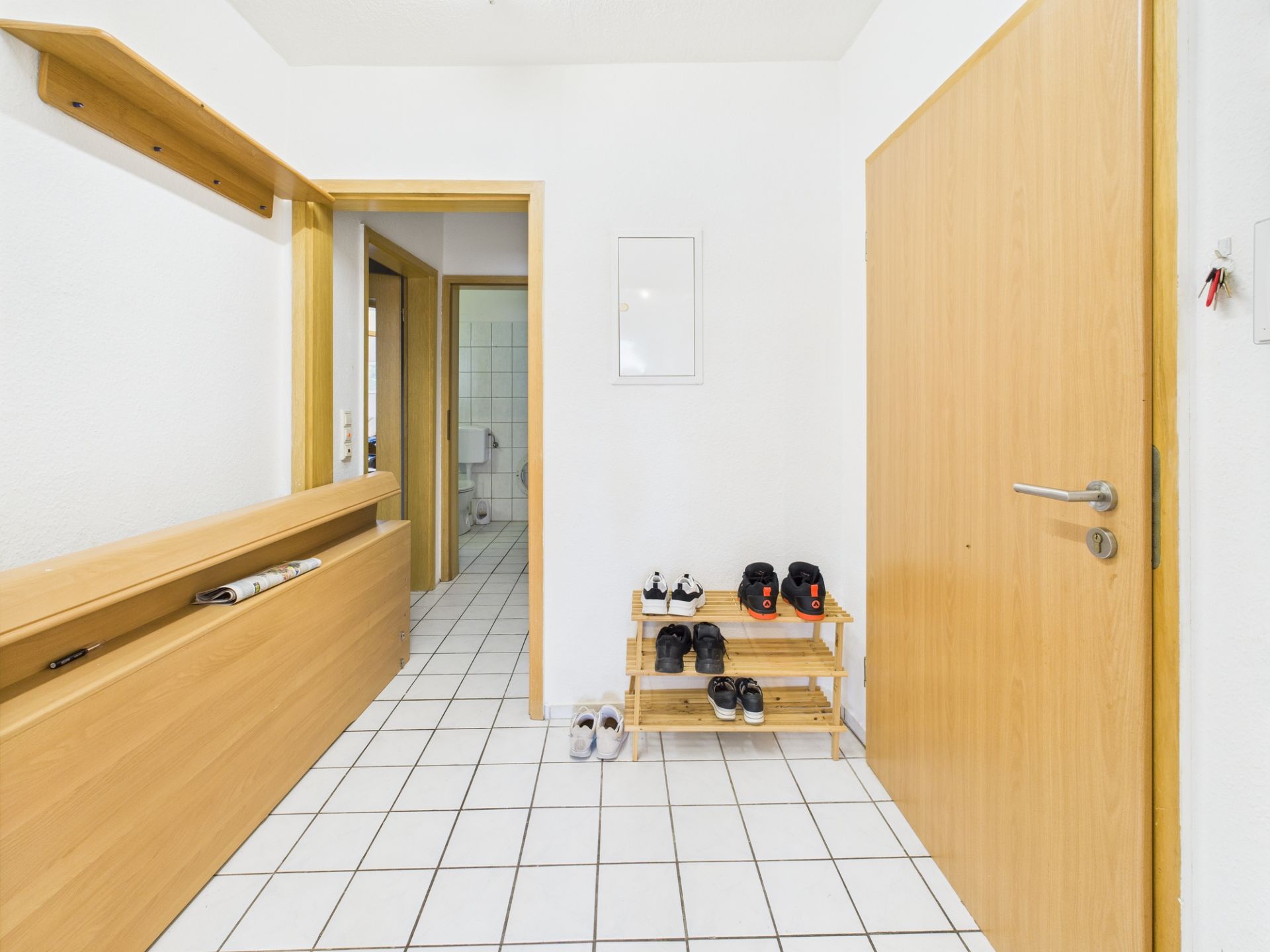 Heller Flur mit weißen Fliesen, Holzregal für Schuhe und offener Tür zu einem Badezimmer.