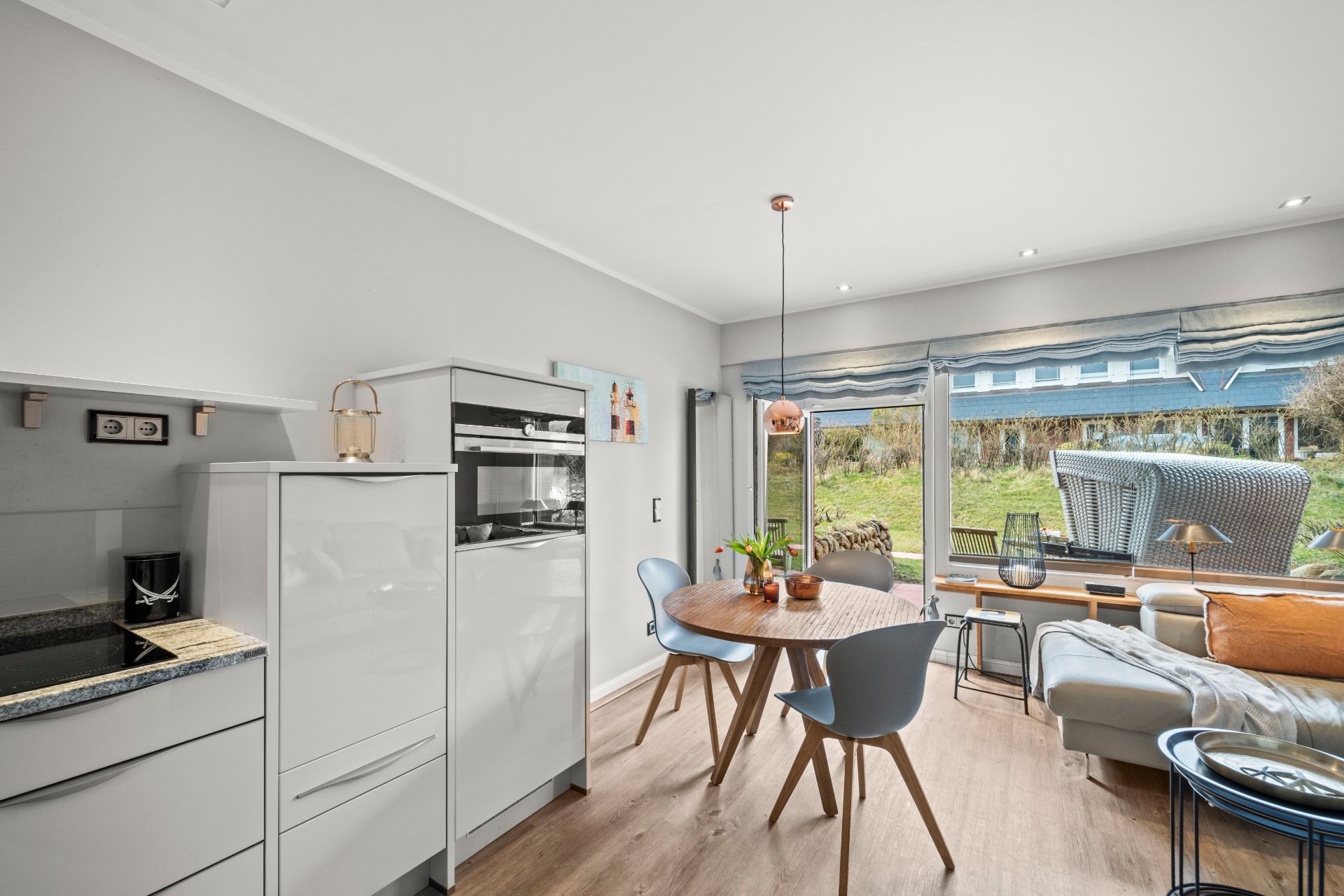 Offene Küche mit Essbereich, rundem Holztisch und Sofa vor großen Fenstern mit Blick auf den Garten.