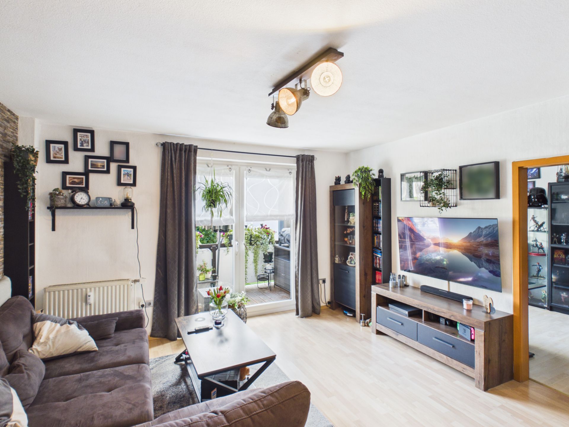 Wohnzimmer mit Sofa, Fernseher, Couchtisch und Zugang zu einem Balkon mit Pflanzen.