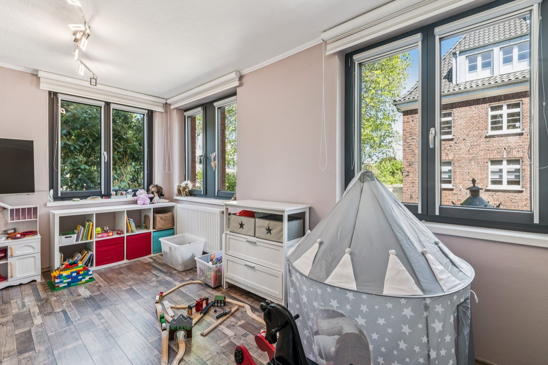 Helles Kinderzimmer mit Spielzeug, Zelt und großen Fenstern, Blick auf Nachbargebäude.
